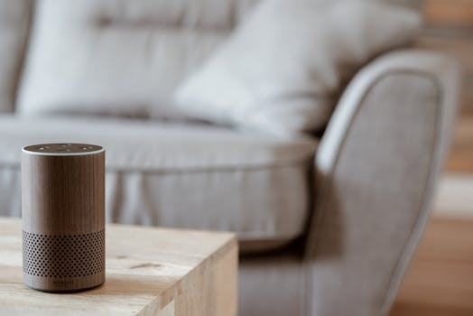 A modern smart speaker sits on a wooden table in a cozy, minimalist living room setting.