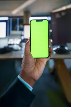 A person holding a smartphone with a green screen in an office setting.