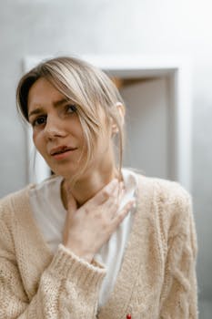 A young woman indoors holds her neck, expressing potential distress or discomfort.