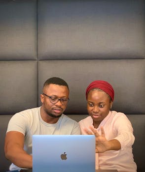 Two people collaborating on a laptop indoors with a modern wall backdrop.