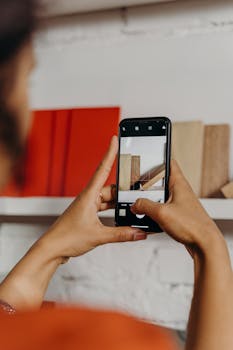 Close-up of hands holding a smartphone to photograph books on a shelf indoors.