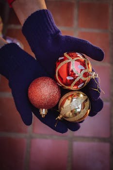 Close-up of hands in gloves holding three colorful Christmas ornaments on a tiled floor.