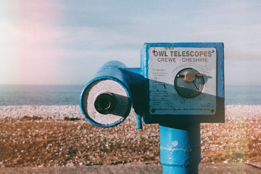 Vintage blue telescope by a pebble beach with ocean view under a clear sky.